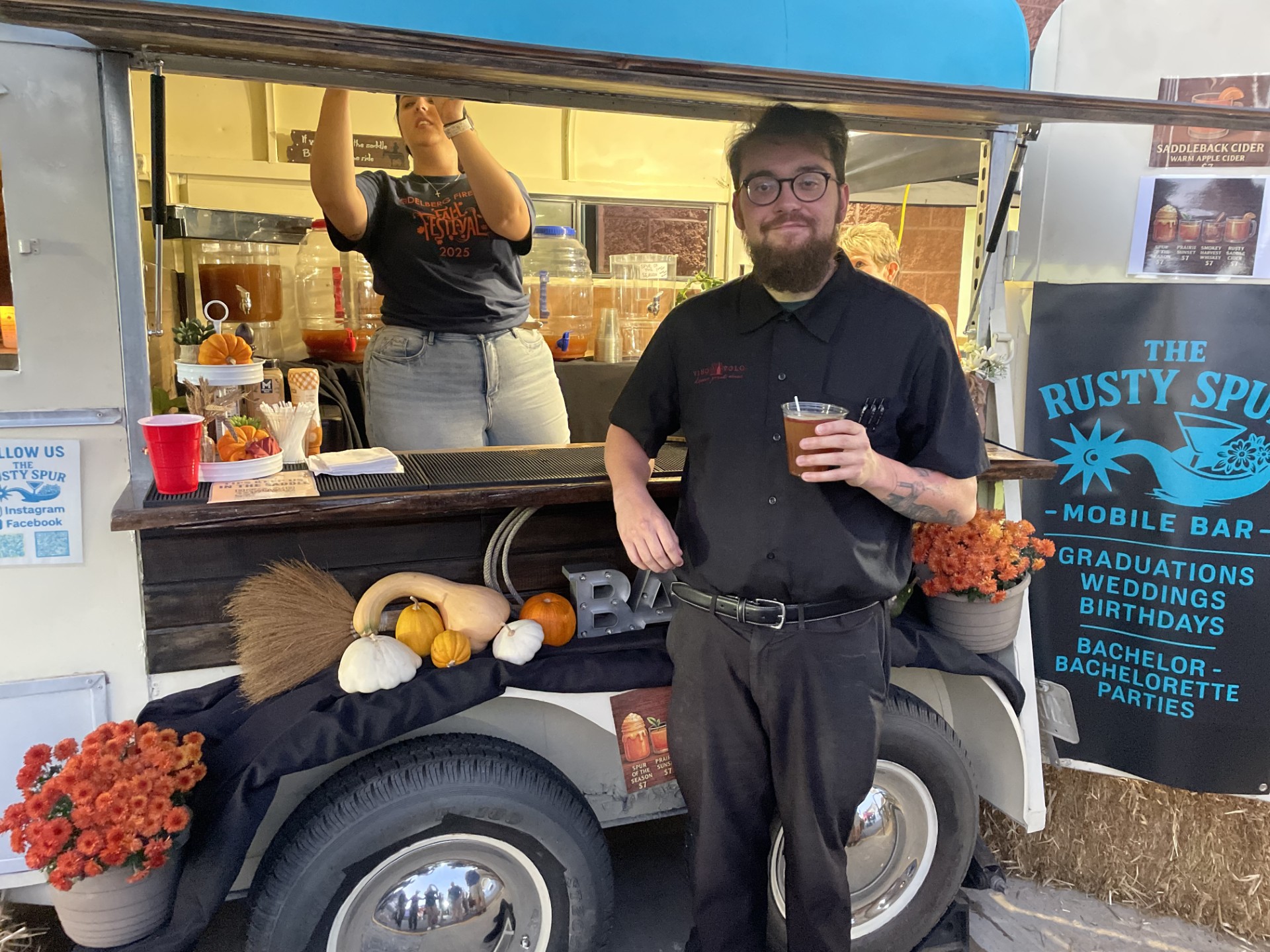 A happy guest with a warm cider in front of the trailer's fall-decorated bar with pumpkins and mums