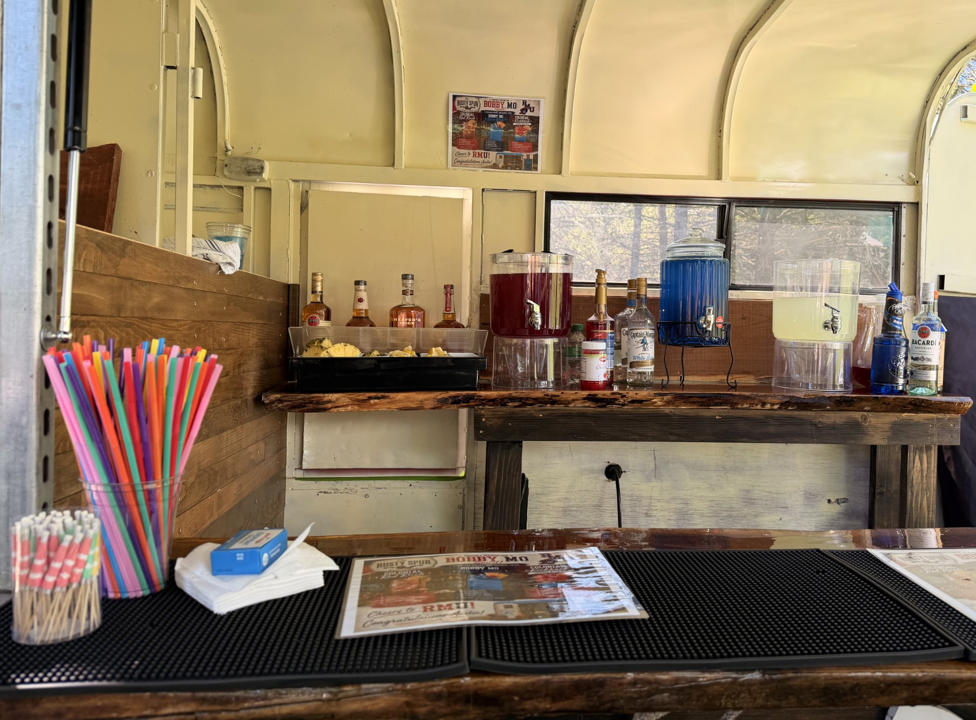 Inside The Rusty Spur trailer — rustic wood bar top stocked with spirits, dispensers, and colorful straws