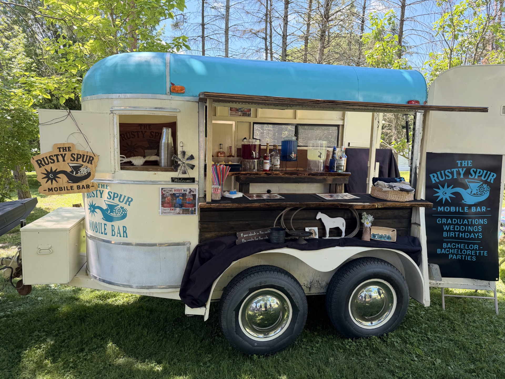 The Rusty Spur cream horse-trailer bar with its turquoise roof fully set up on a green lawn, chalkboard sign out front
