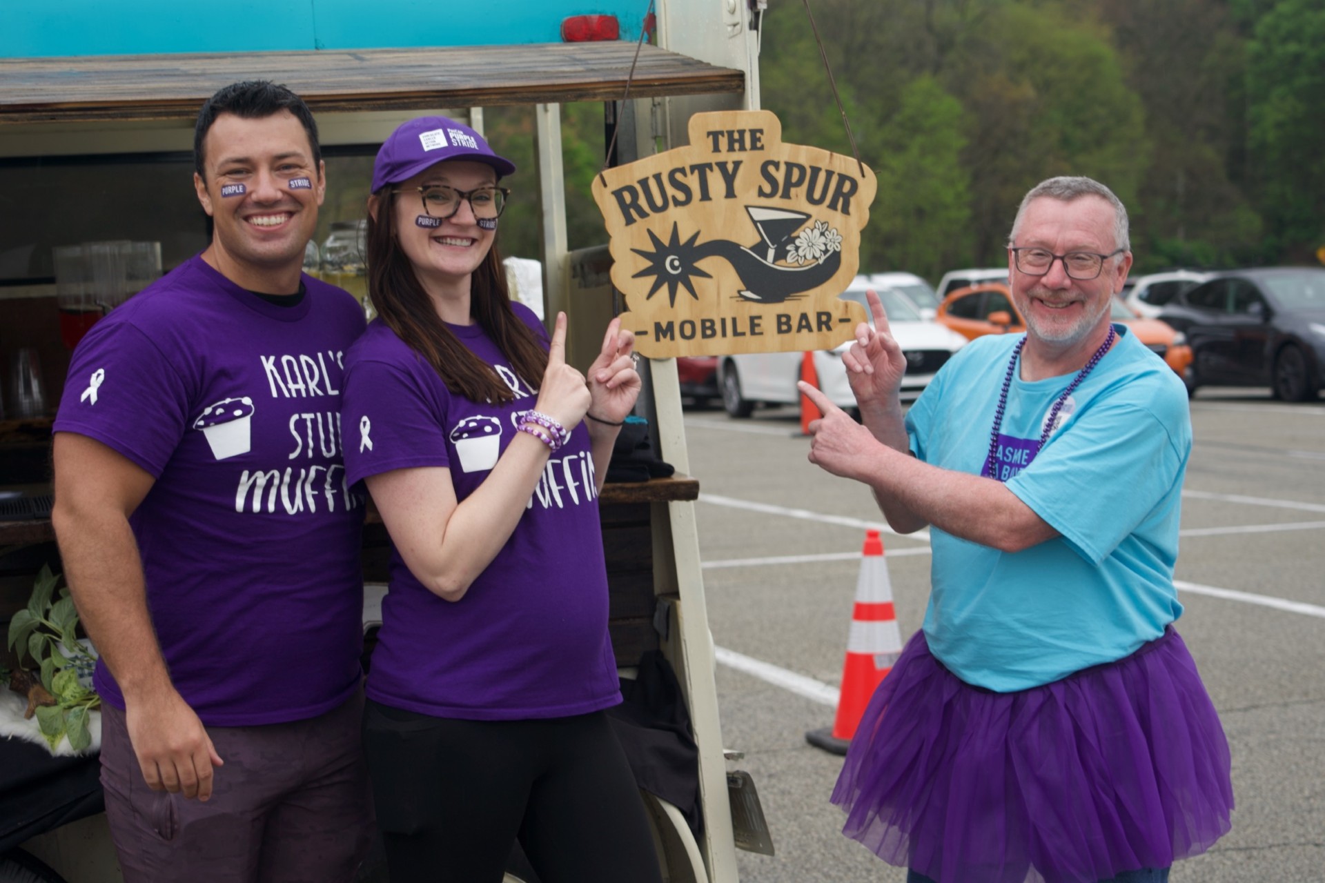 Three friends pointing at the hand-painted wooden Rusty Spur sign at a charity event