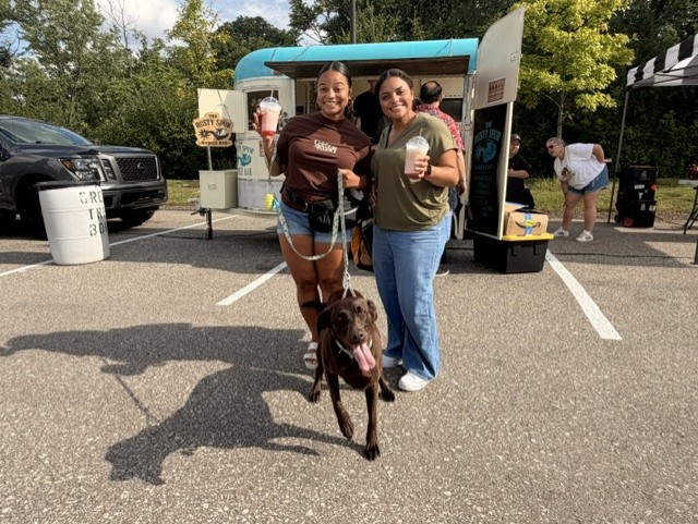 Two guests holding drinks in front of The Rusty Spur trailer with their chocolate lab at an outdoor event